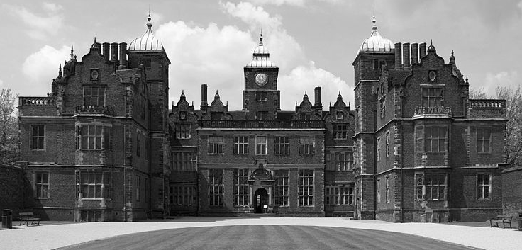 A grand, imposing brick manor house with clock tower, under a cloudy sky.