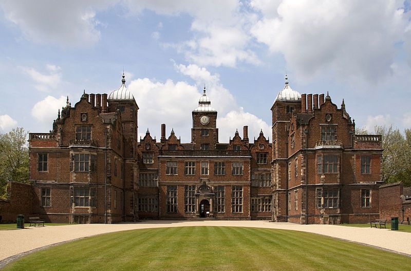 Large brick manor house with ornate towers and clock, on a grassy lawn under a sunny sky.