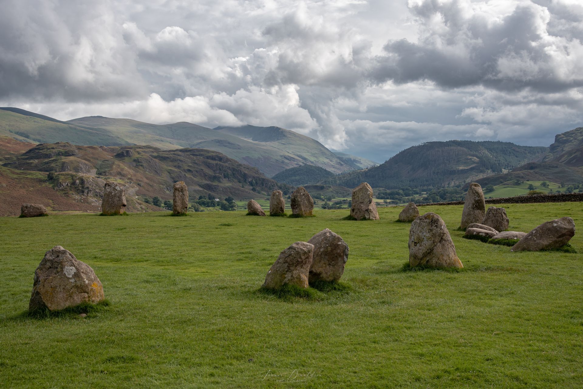 Stone circle in a green field, with hills and cloudy sky in the background.