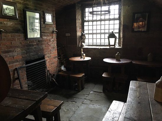 A dimly lit, rustic tavern interior with brick walls, small round tables, and a barred window.