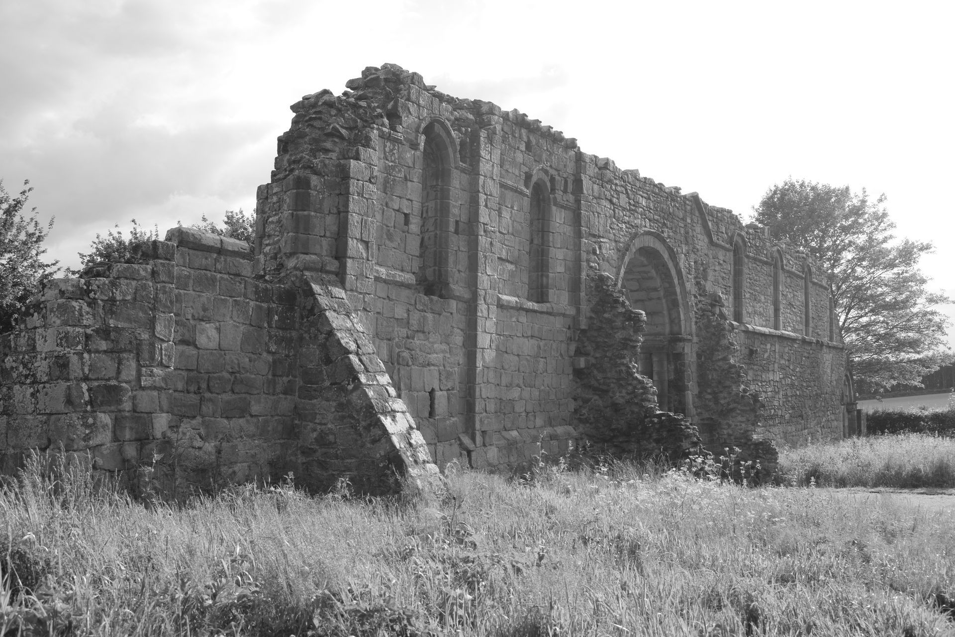 Ruins of stone building, overgrown with grass and vegetation, under cloudy sky.