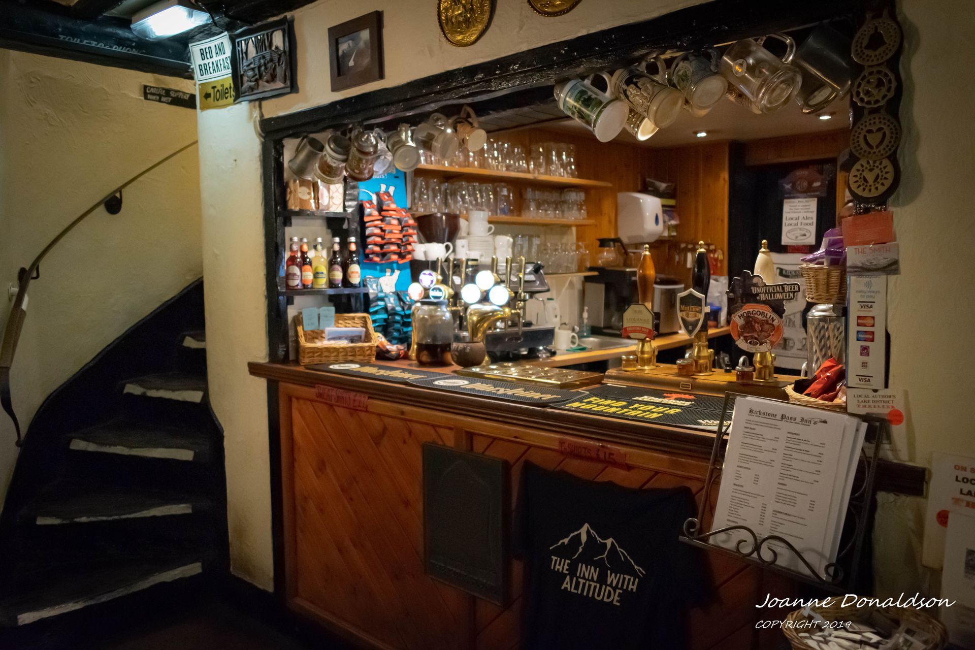 Wooden bar inside a pub, with beer taps, glasses, and a staircase to the left.