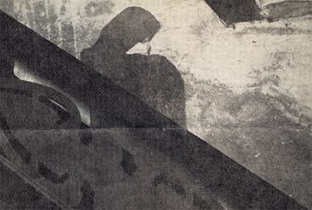 Shadow of a person with a hood cast on a textured wall, with dark diagonal beams in the foreground.
