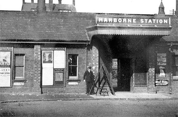 Exterior of Harborne Station. A man stands near the entrance under a canopy. Posters visible, brick building.