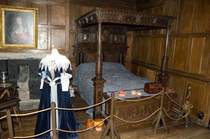 Bedroom with a four-poster bed, fireplace, portrait, and blue gown on a mannequin, inside a historic building.