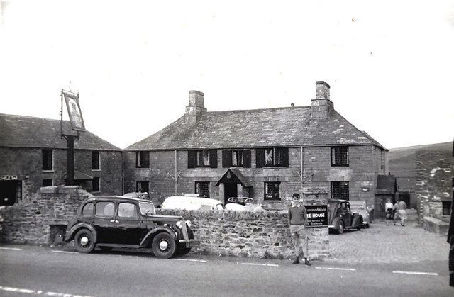 A vintage black and white photo of a stone pub with parked cars. A sign hangs outside.