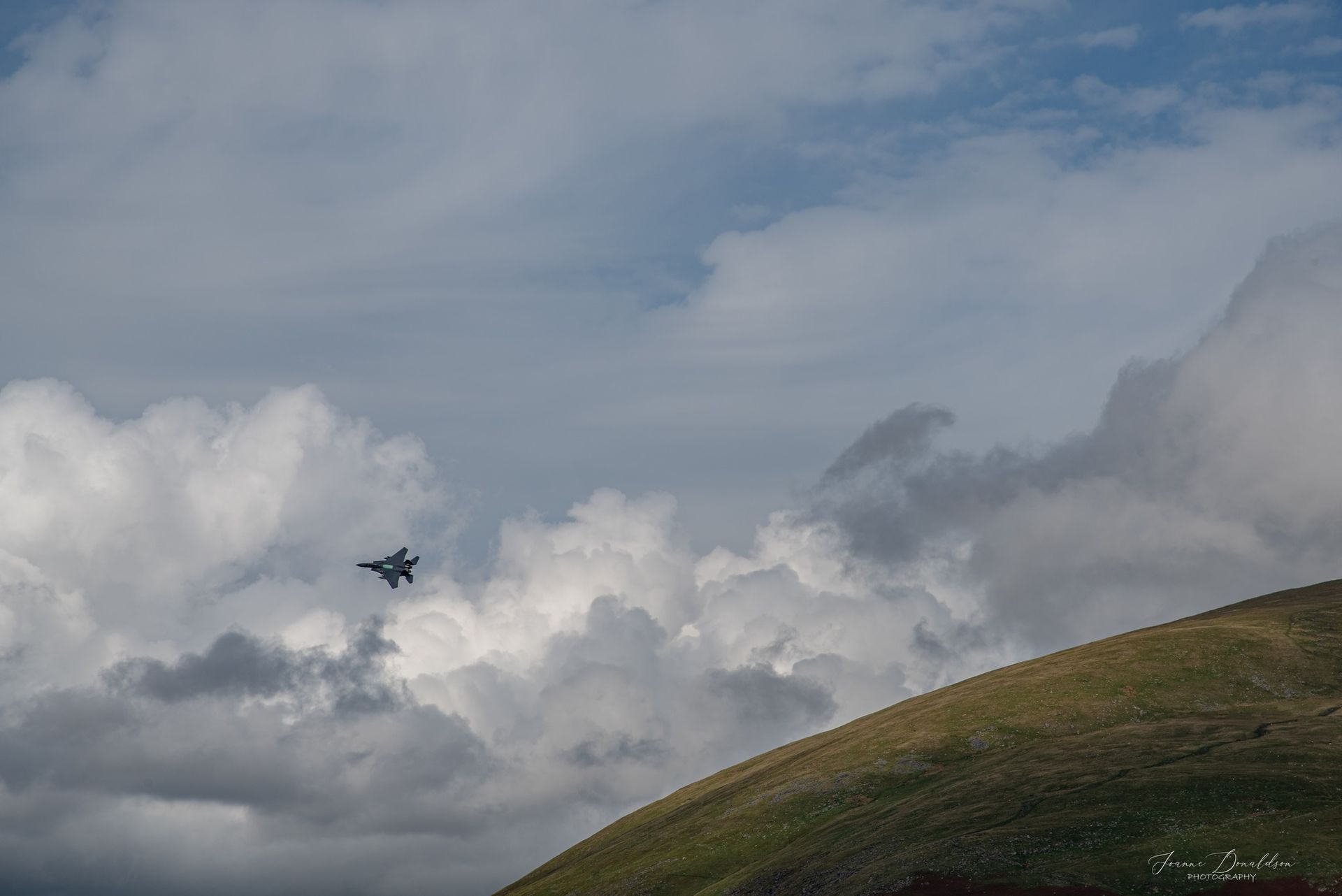 Jet fighter in flight, against a cloudy sky, near a green hillside.