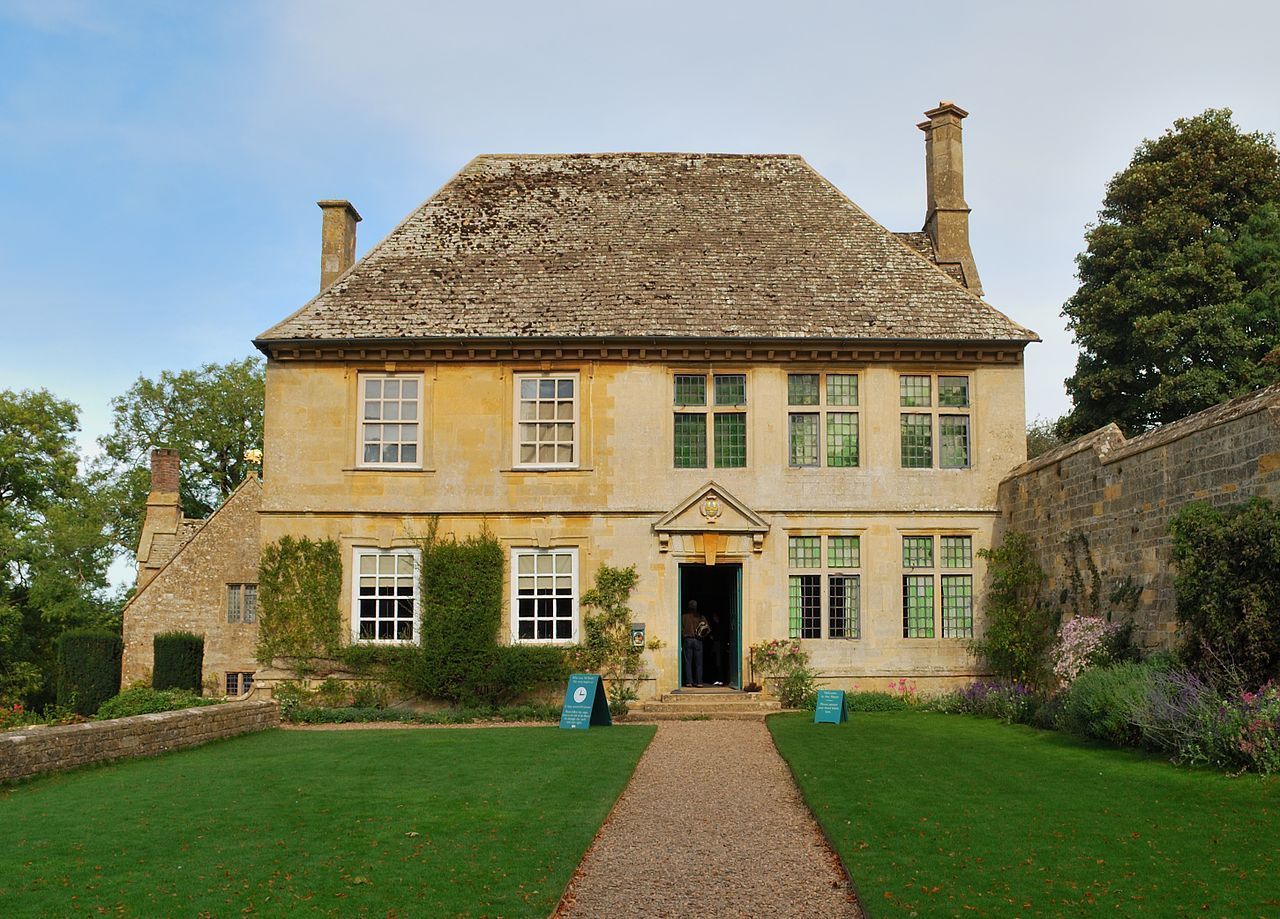 A stone-built manor house with a mossy roof and green lawn under a cloudy sky.