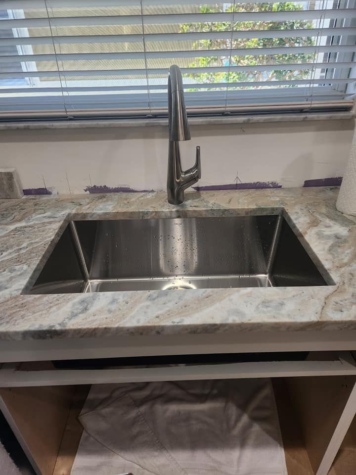 A stainless steel kitchen sink with a tall chrome faucet, installed in a light-colored granite countertop.
