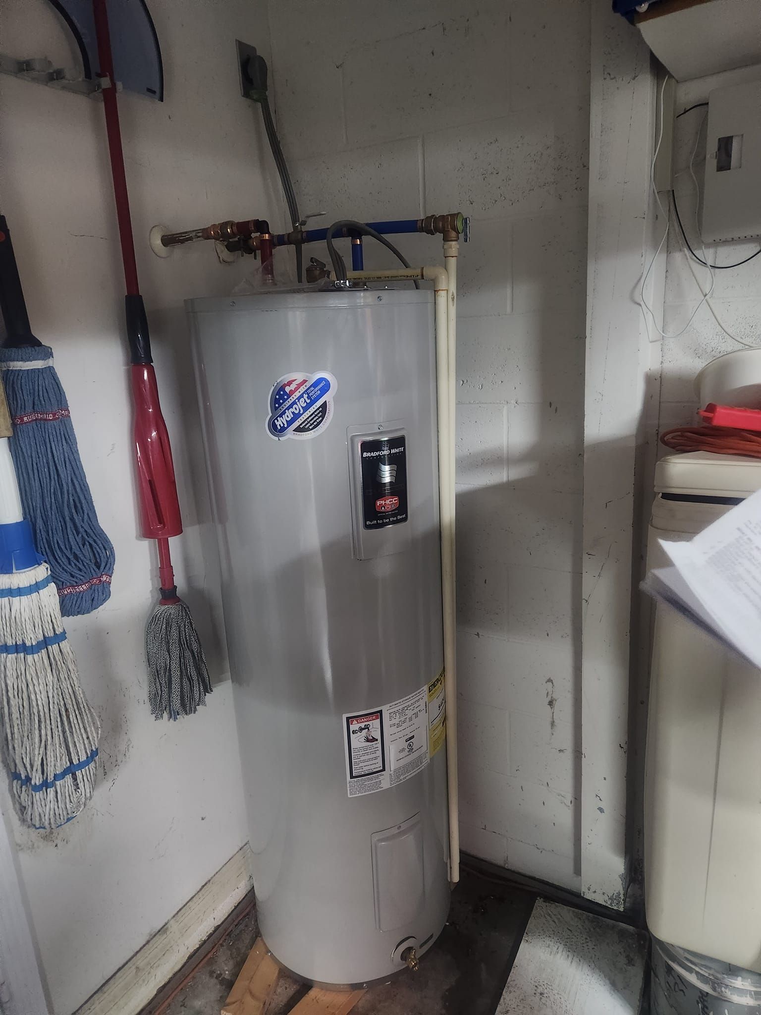 A light gray electric water heater stands in a corner with plumbing connections on top and cleaning mops hanging nearby.
