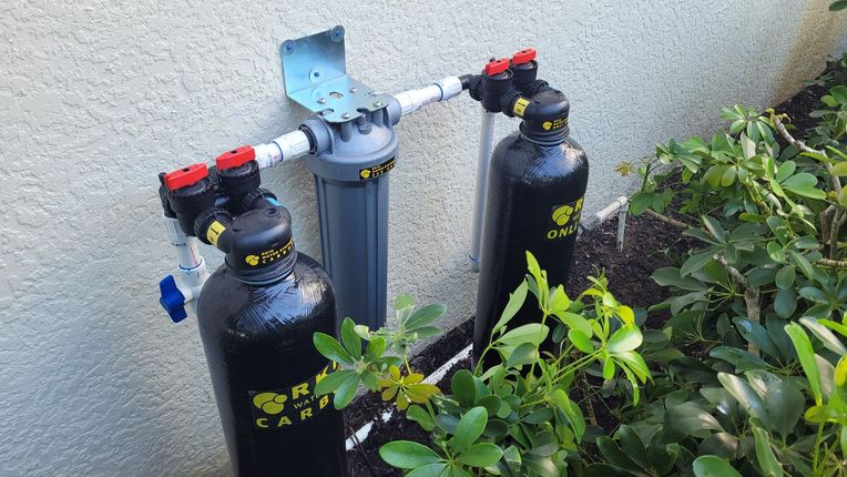 A whole-house water filtration system with two black tanks and a central gray filter housing mounted on an outdoor wall.