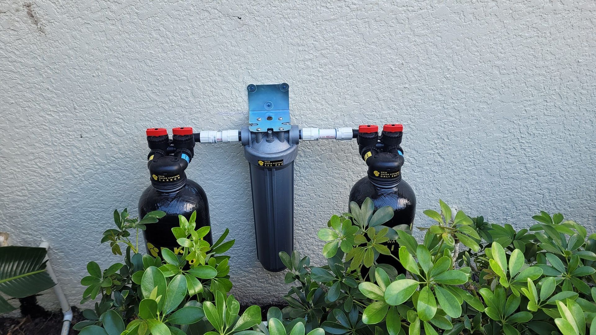 A water filtration system with two black tanks and a central grey housing mounted on a beige stucco wall behind shrubs.