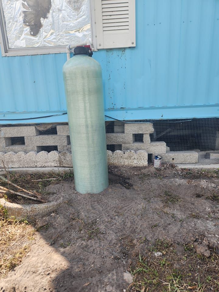 A light green, cylindrical water filtration tank stands on bare dirt outside a blue mobile home near a cinderblock skirt.