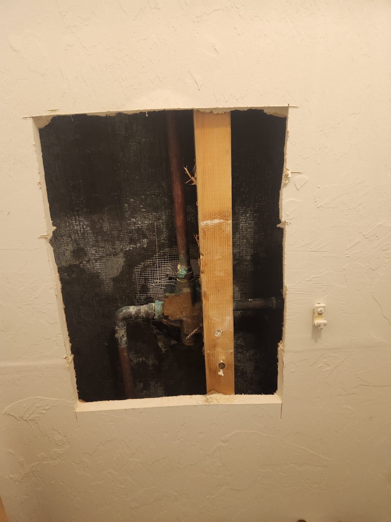 A rectangular hole cut into a white textured wall reveals plumbing pipes and a wooden stud behind the drywall.