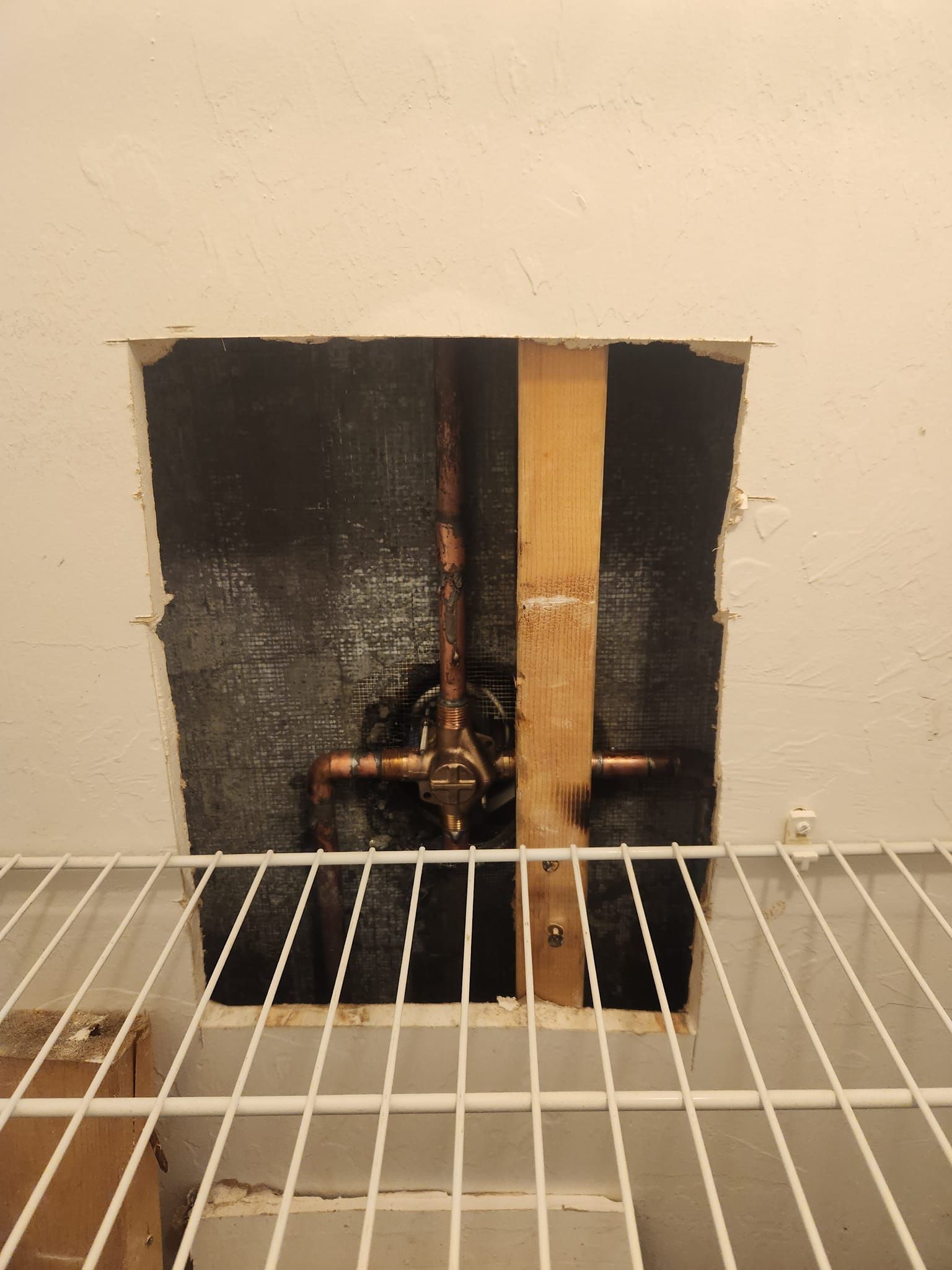 A square hole cut into a white wall reveals copper plumbing pipes and a wooden stud behind a wire shelving unit.
