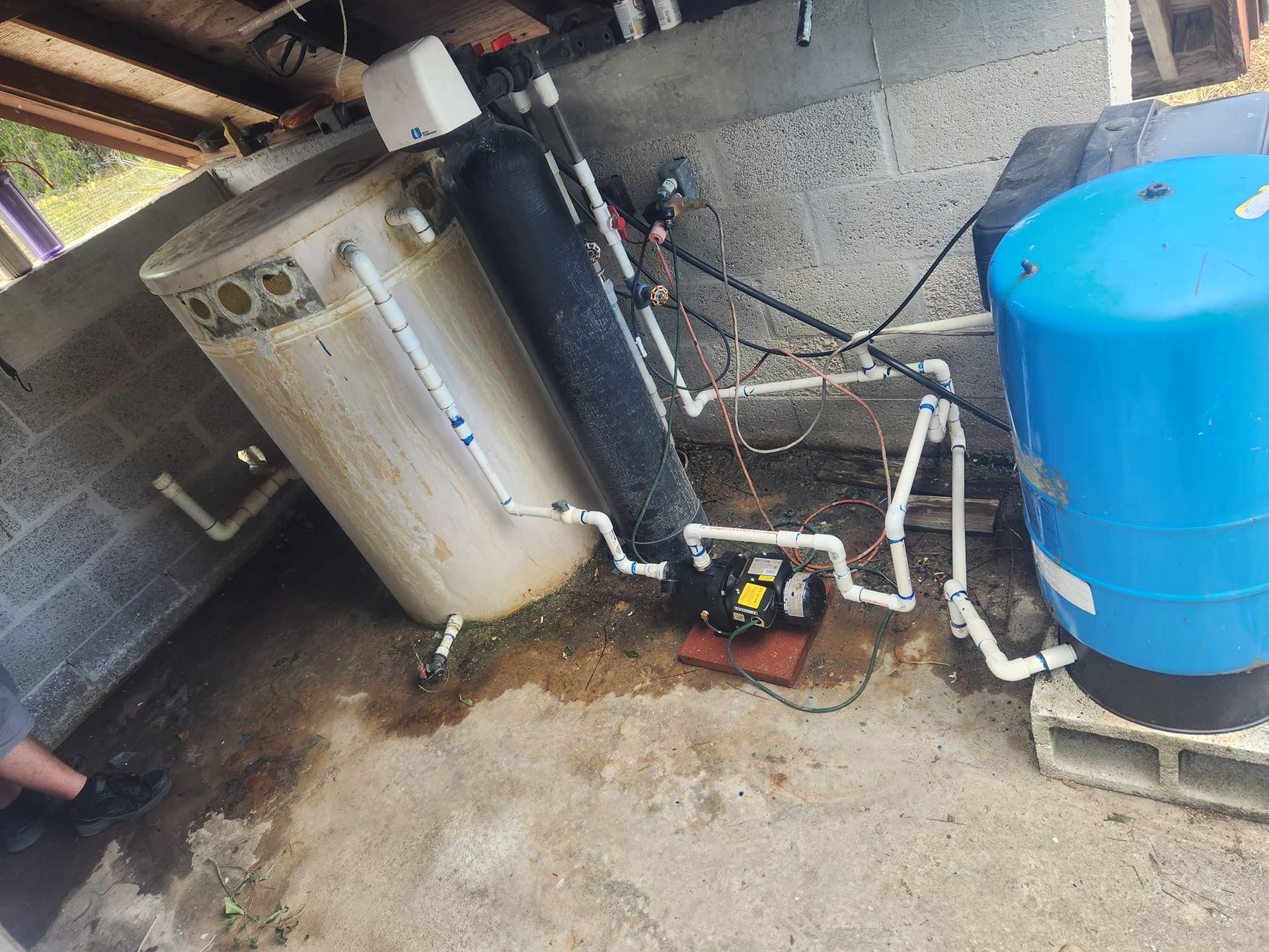 A water well system in a garage featuring a large white holding tank, a black filter cylinder, a pump, and a blue tank.