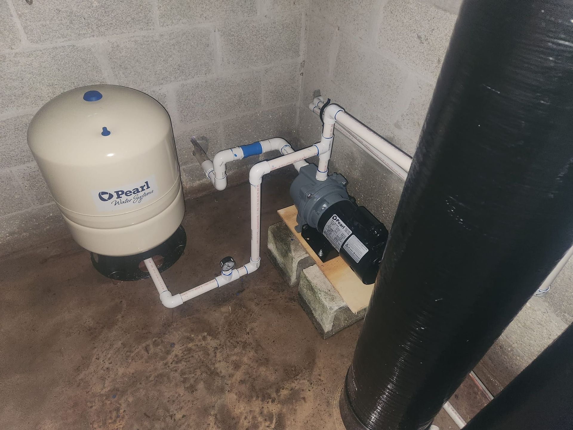 A beige pressure tank connected by white PVC pipes to a black water pump resting on a concrete block in a basement corner.