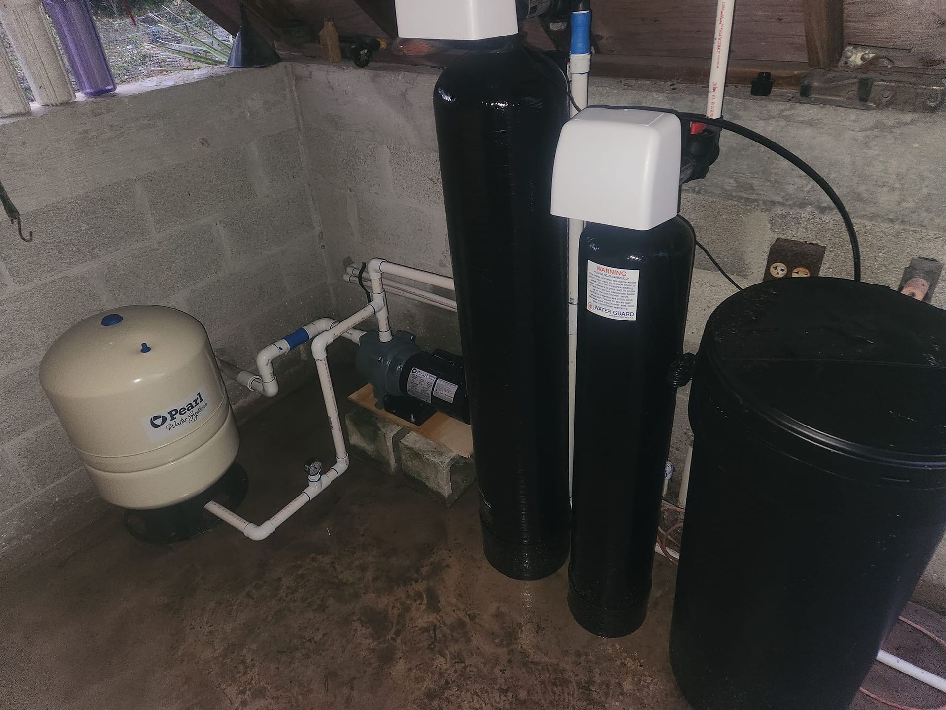 Water filtration system with a beige pressure tank, two black tanks, and a pump against a cinder block wall.