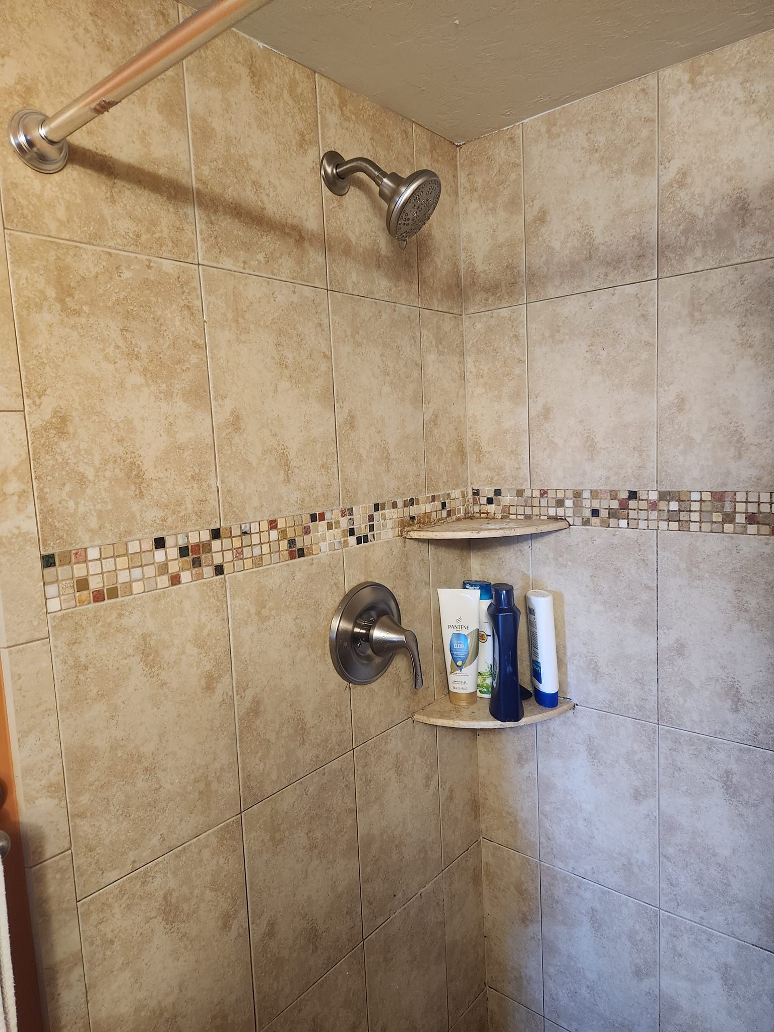 A shower stall featuring tan stone-look tiles, a chrome showerhead, a single handle mixer, and corner storage shelves.