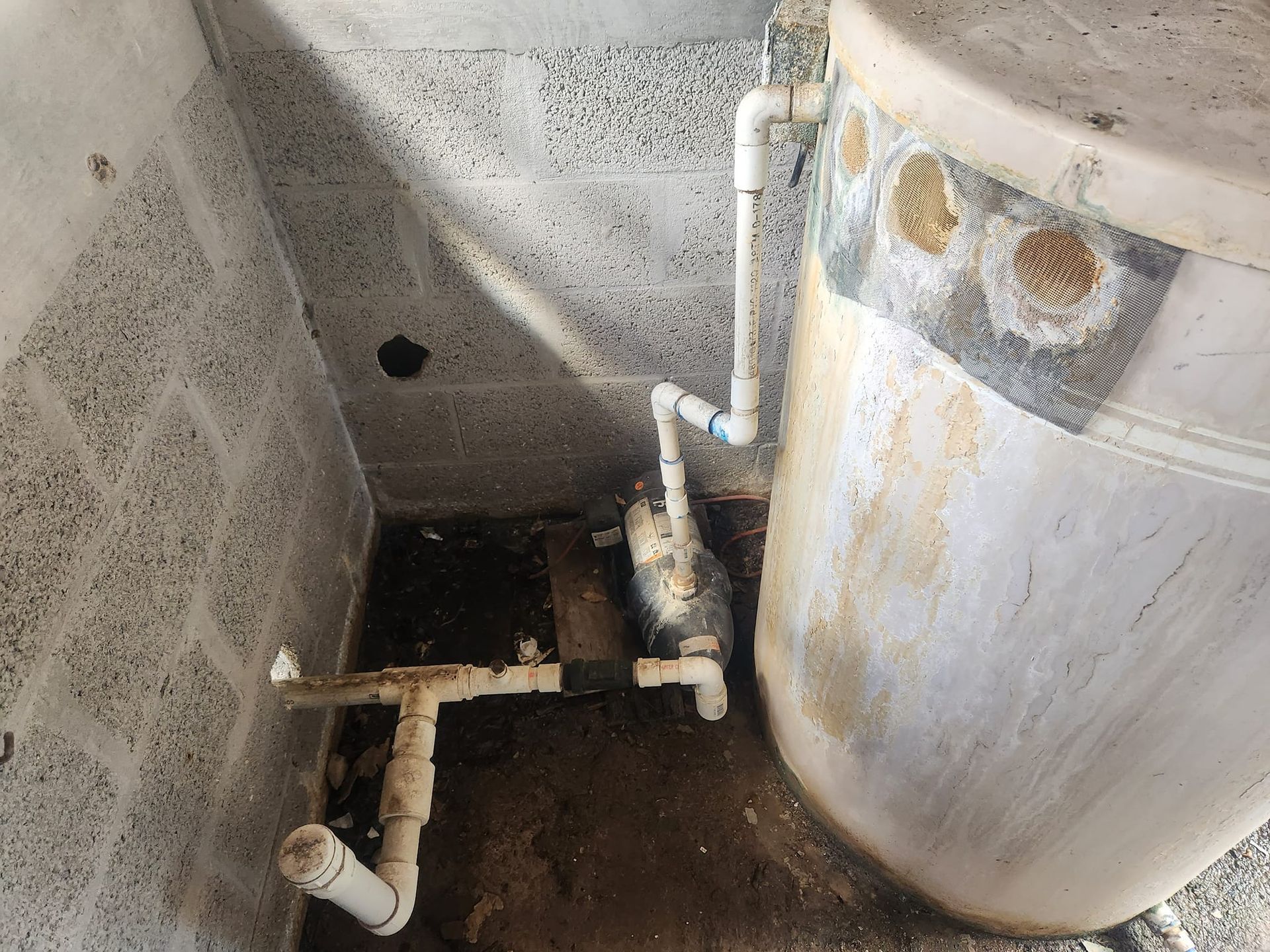 White PVC piping connects a water tank to a floor-mounted pump in a basement with concrete block walls.