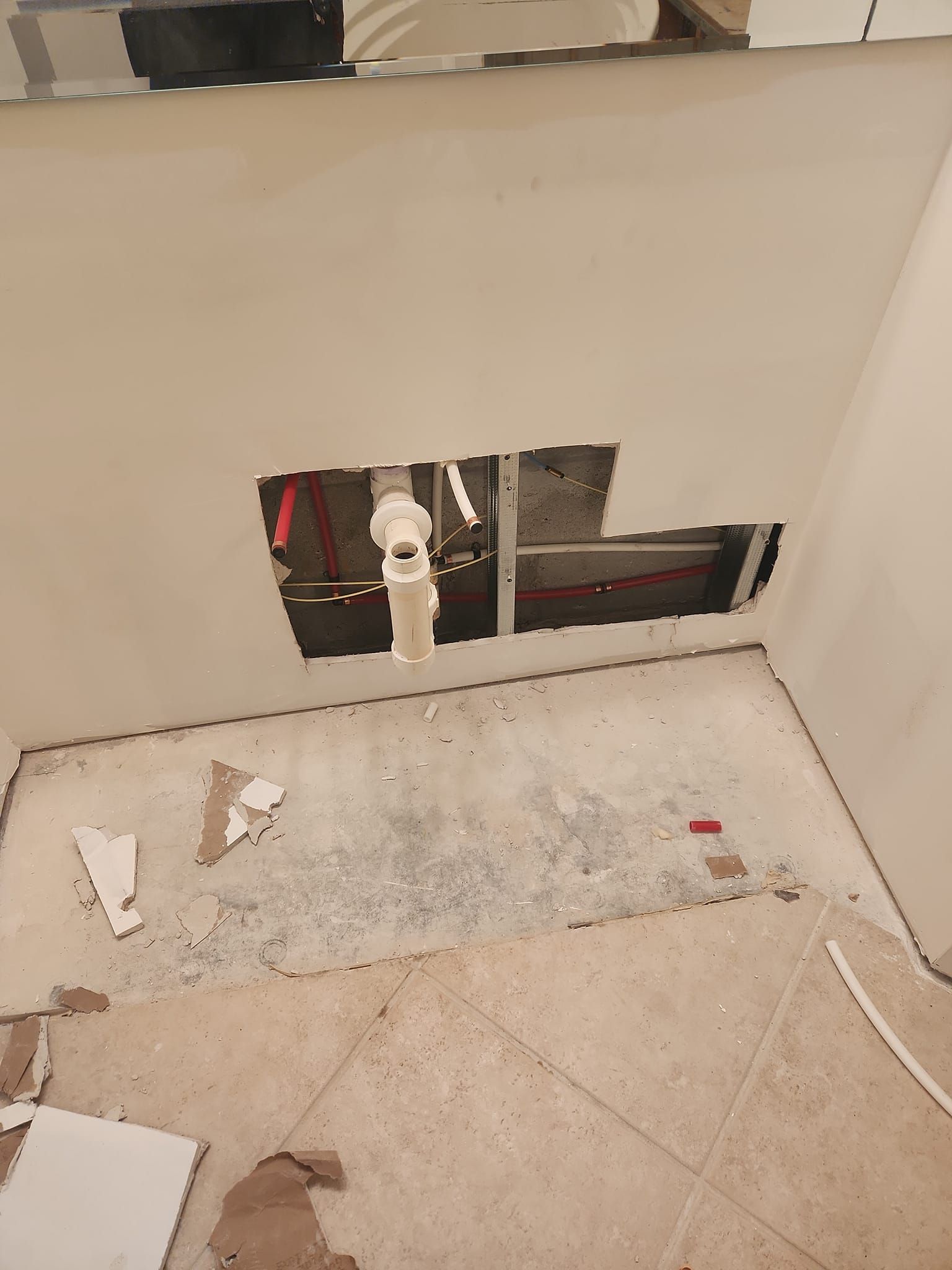 A large rectangular hole cut into a beige wall, exposing wall studs and plumbing pipes above a tiled bathroom floor.