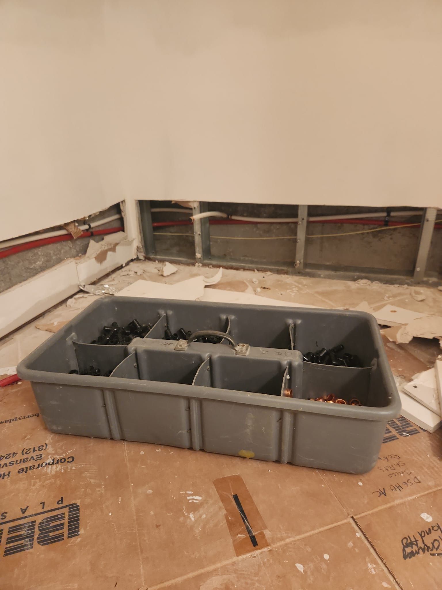 A grey plastic utility bin sits on a construction site floor in front of a wall with exposed metal studs and pipes.