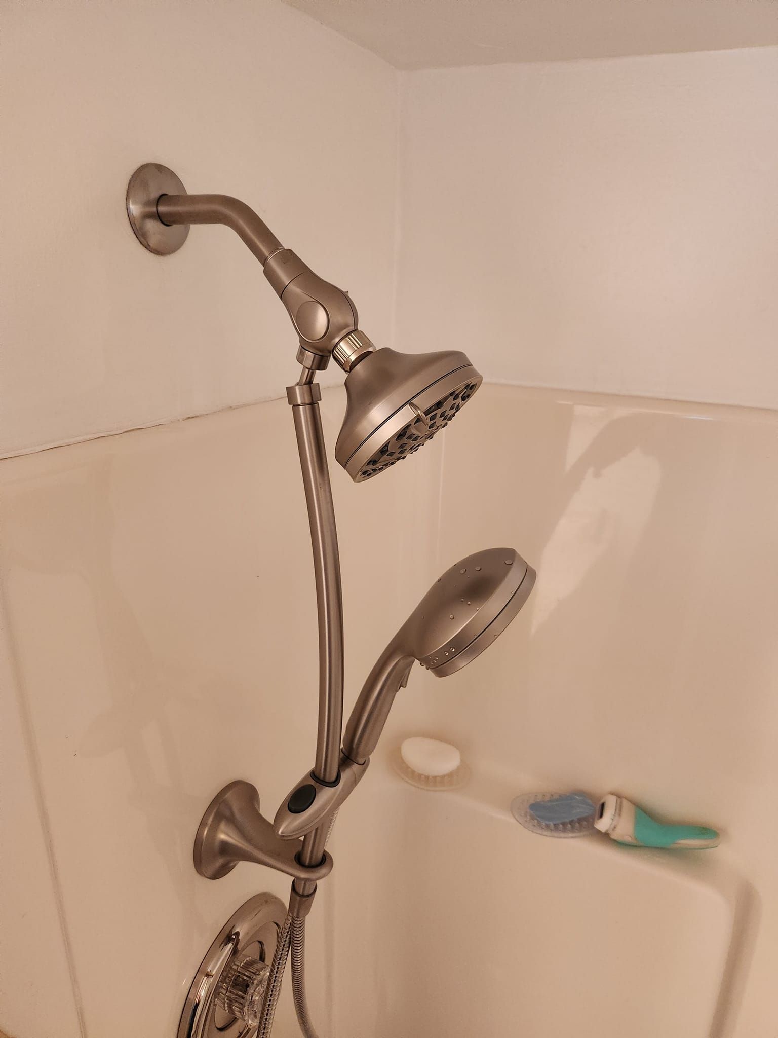 A brushed nickel dual showerhead system with a fixed head and a detachable handheld sprayer in a white shower stall.