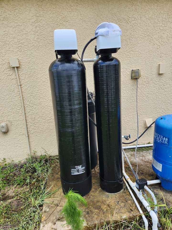 Two tall, black water filtration tanks with white control heads, standing outdoors on a concrete pad near a blue tank.