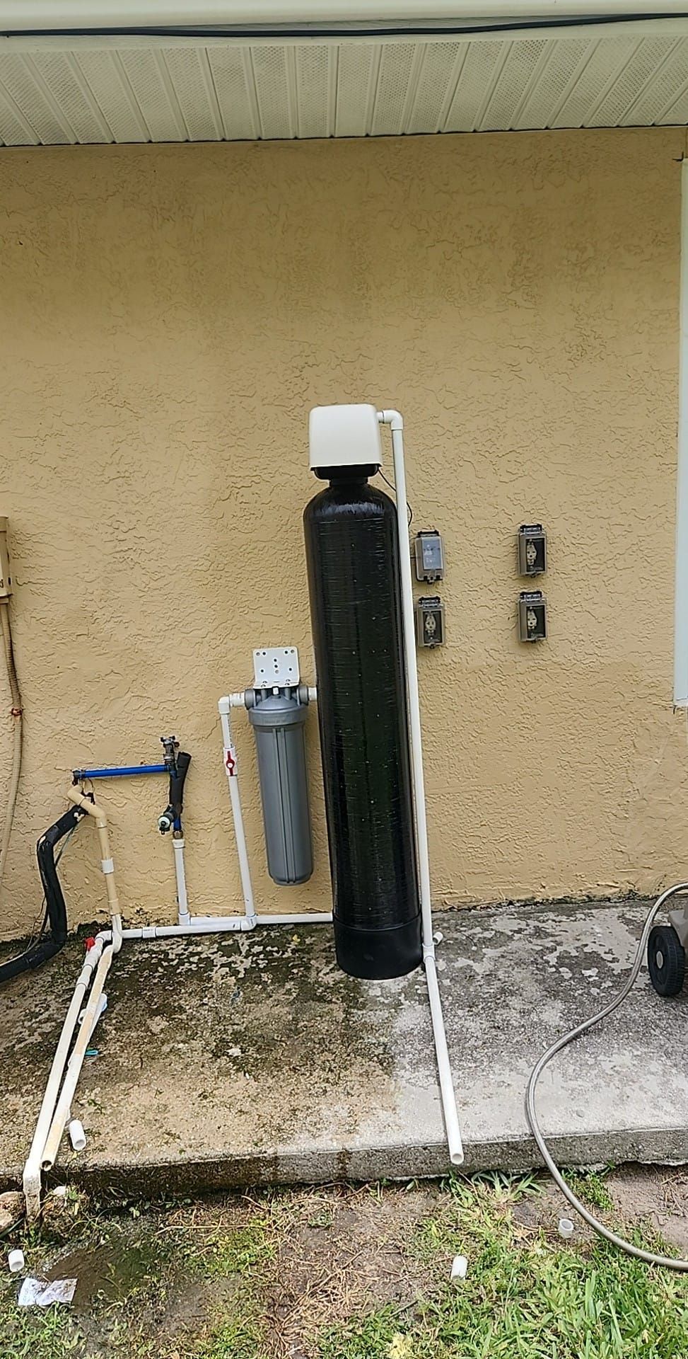 A tall, black water filtration tank and a smaller cylindrical filter housing connected by white PVC pipes outdoors.
