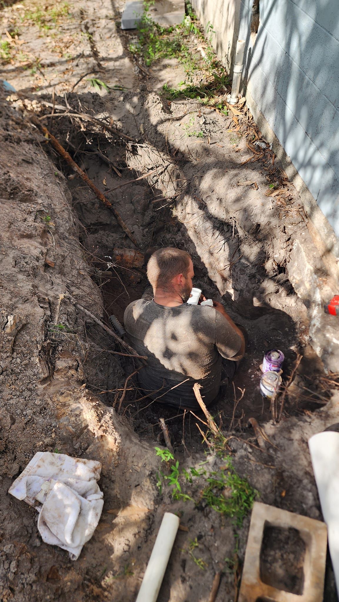 A person crouches inside a narrow trench in the dirt next to a concrete wall while working on plumbing pipes.