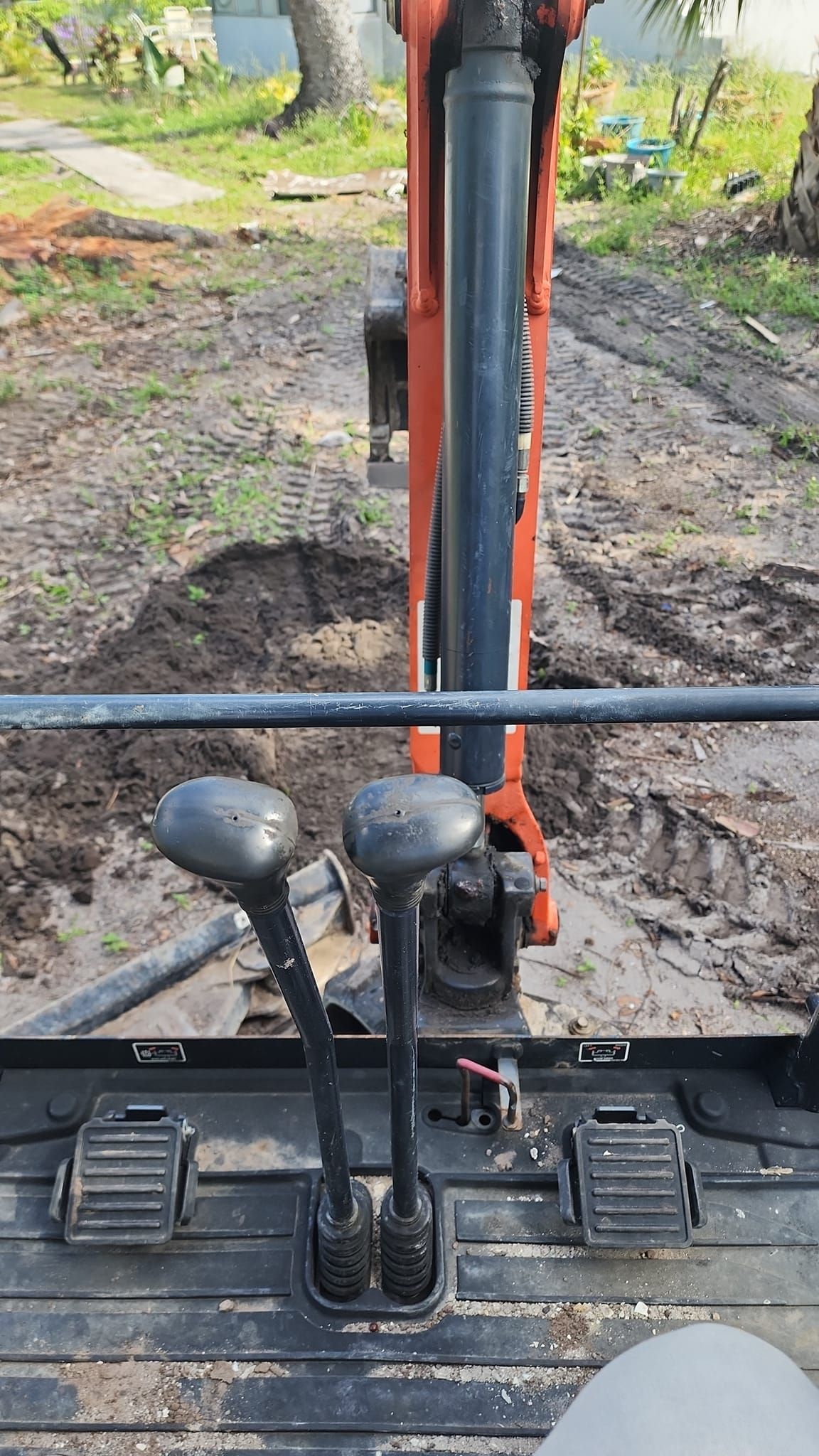 A first-person view from inside an orange excavator, showing the joysticks, foot pedals, and a muddy outdoor work site.