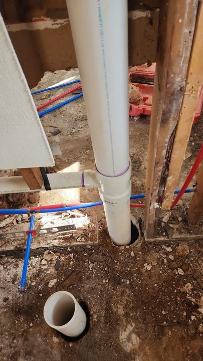 A vertical white PVC pipe extends through the floor, near a wooden wall stud and exposed red and blue PEX water lines.