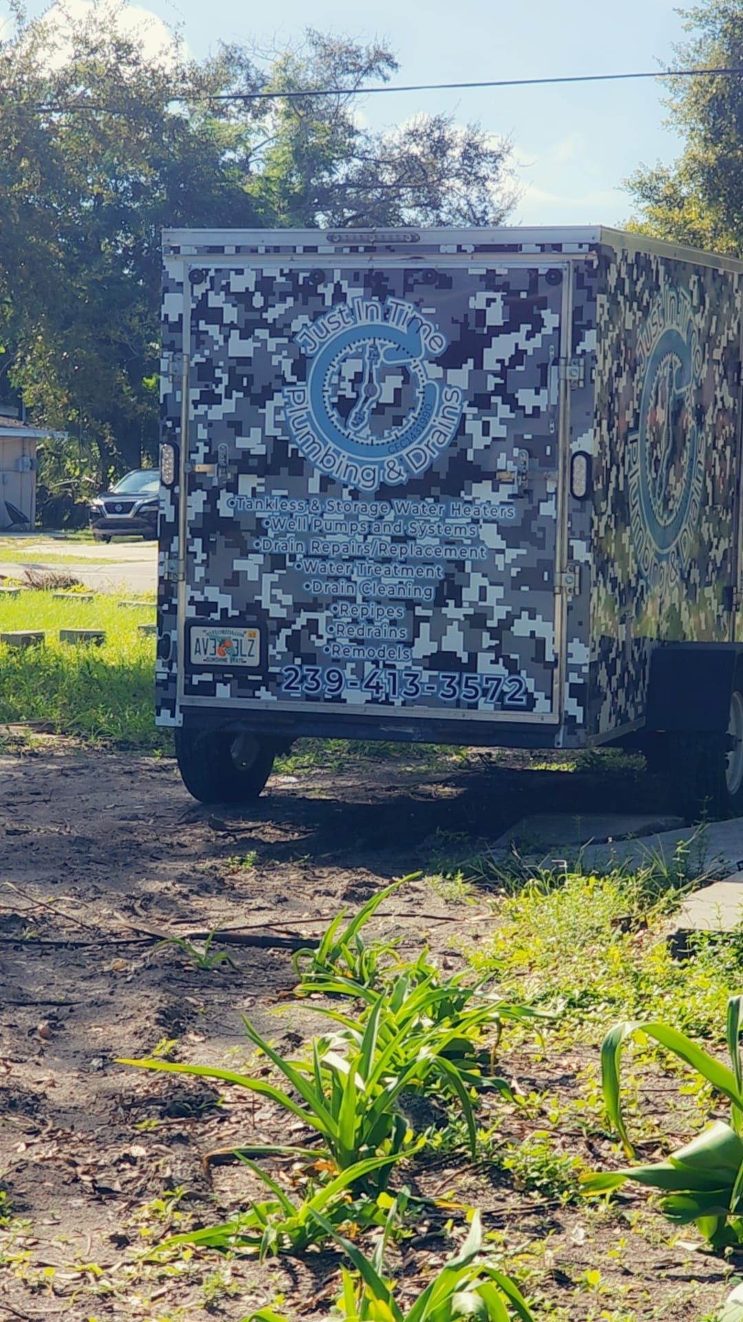 A grey camouflage-patterned utility trailer parked on grass with a blue and white company logo on the rear door.