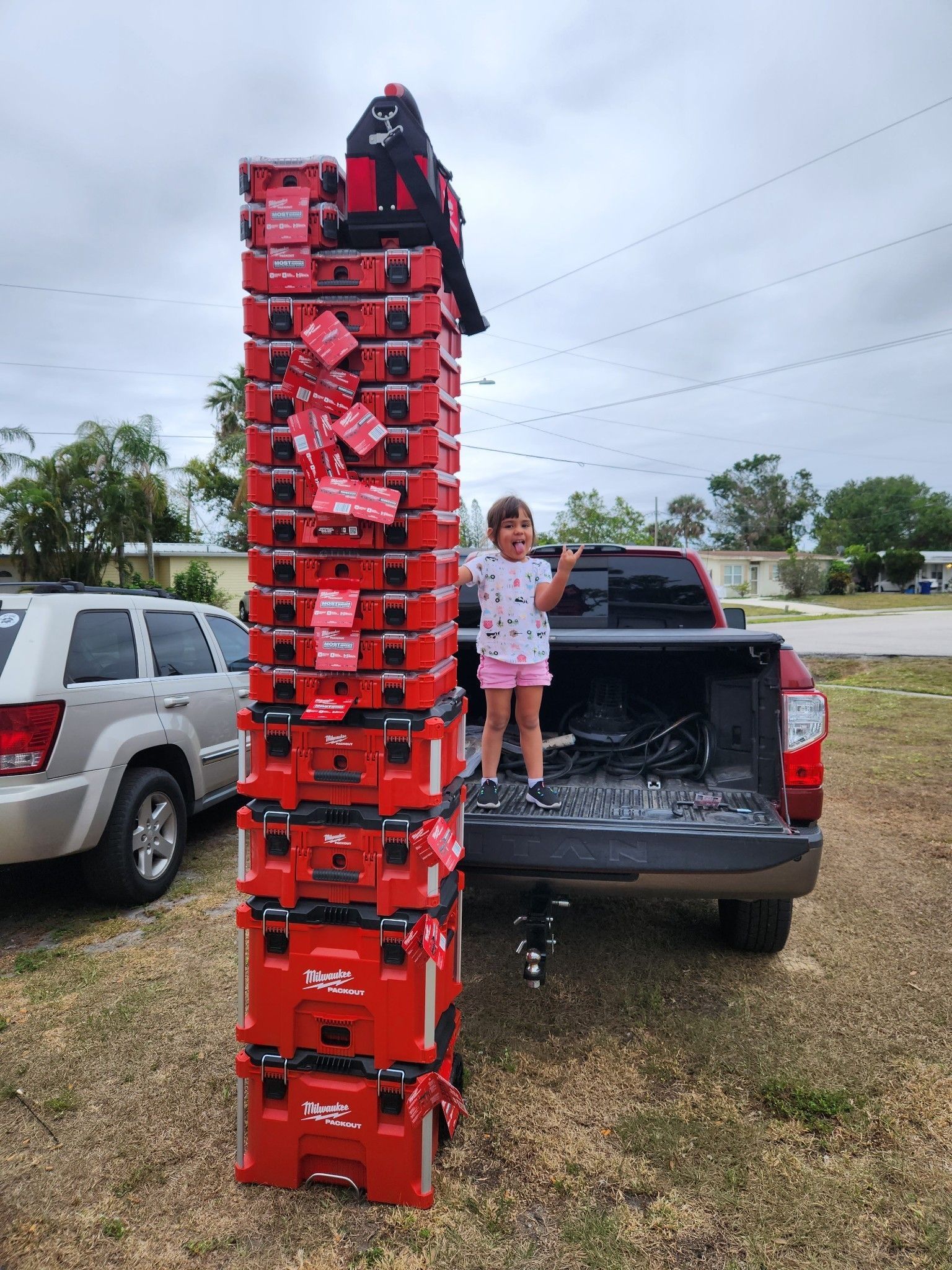 A young child stands next to a massive, precarious vertical stack of red Milwaukee Packout tool cases in a truck bed.