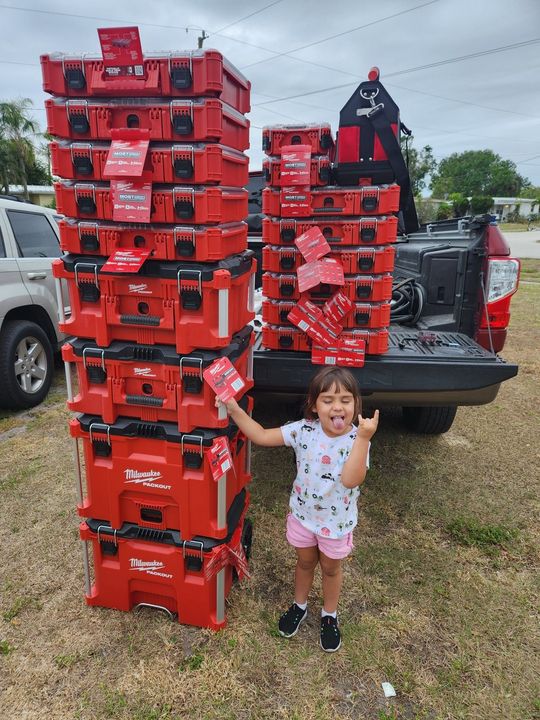 A child playfully makes a rock-on gesture next to two tall stacks of red Milwaukee Packout tool cases in a truck bed.