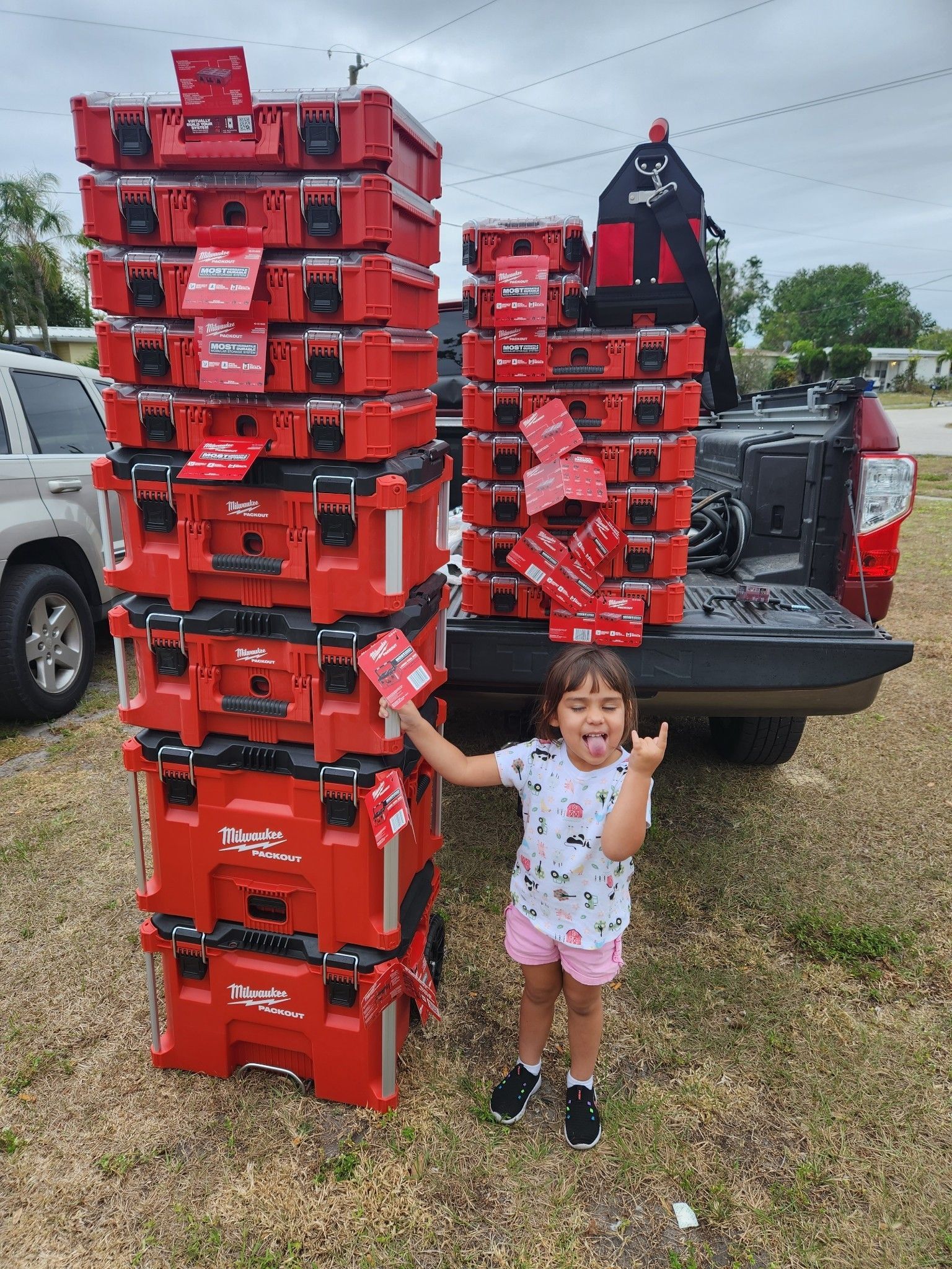 A child poses with a tongue-out, rock-on hand gesture next to two tall stacks of red Milwaukee Packout tool cases.