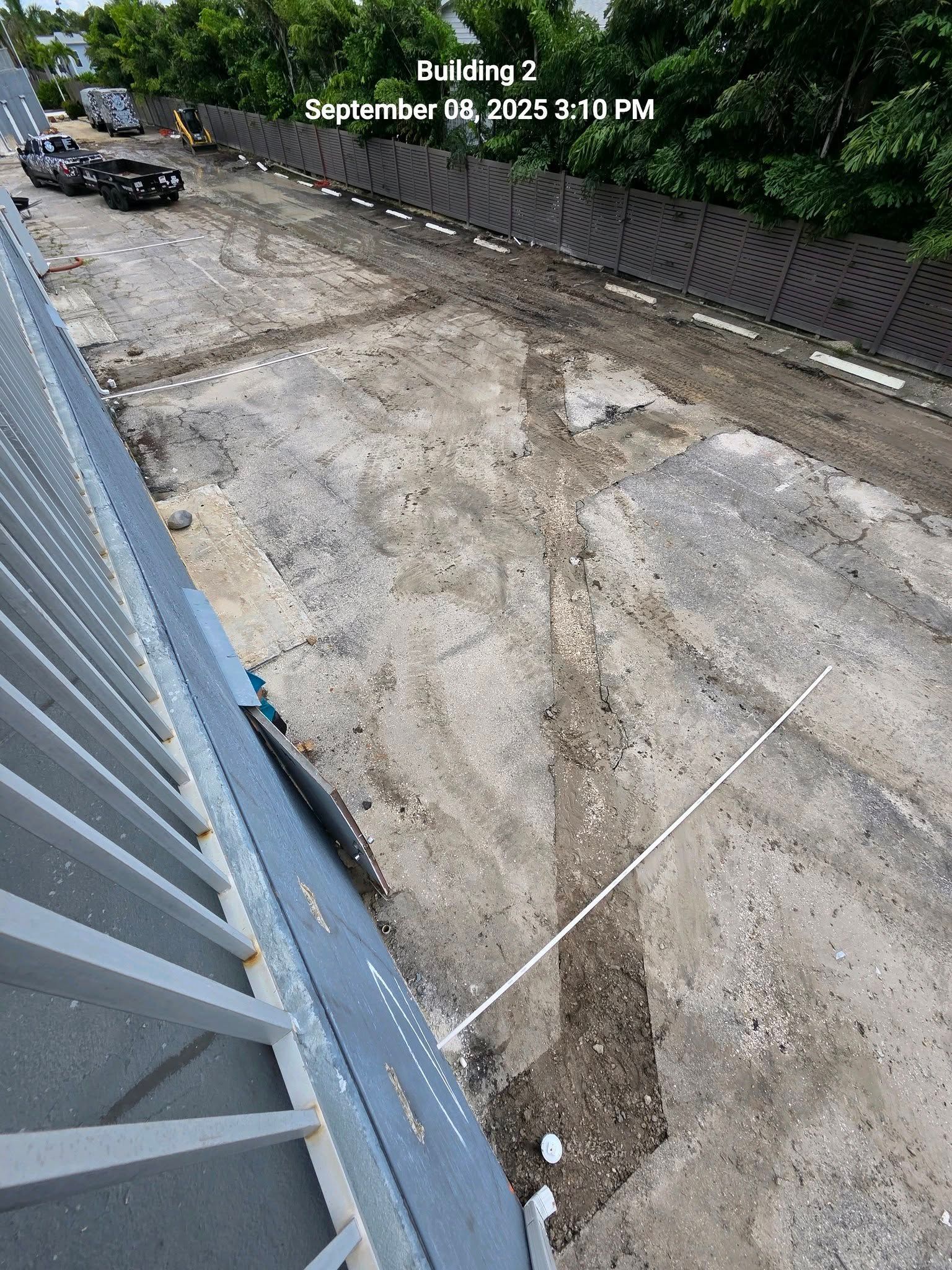 A view from above showing a dirt construction site with tire tracks near a gray building balcony and a brown fence.
