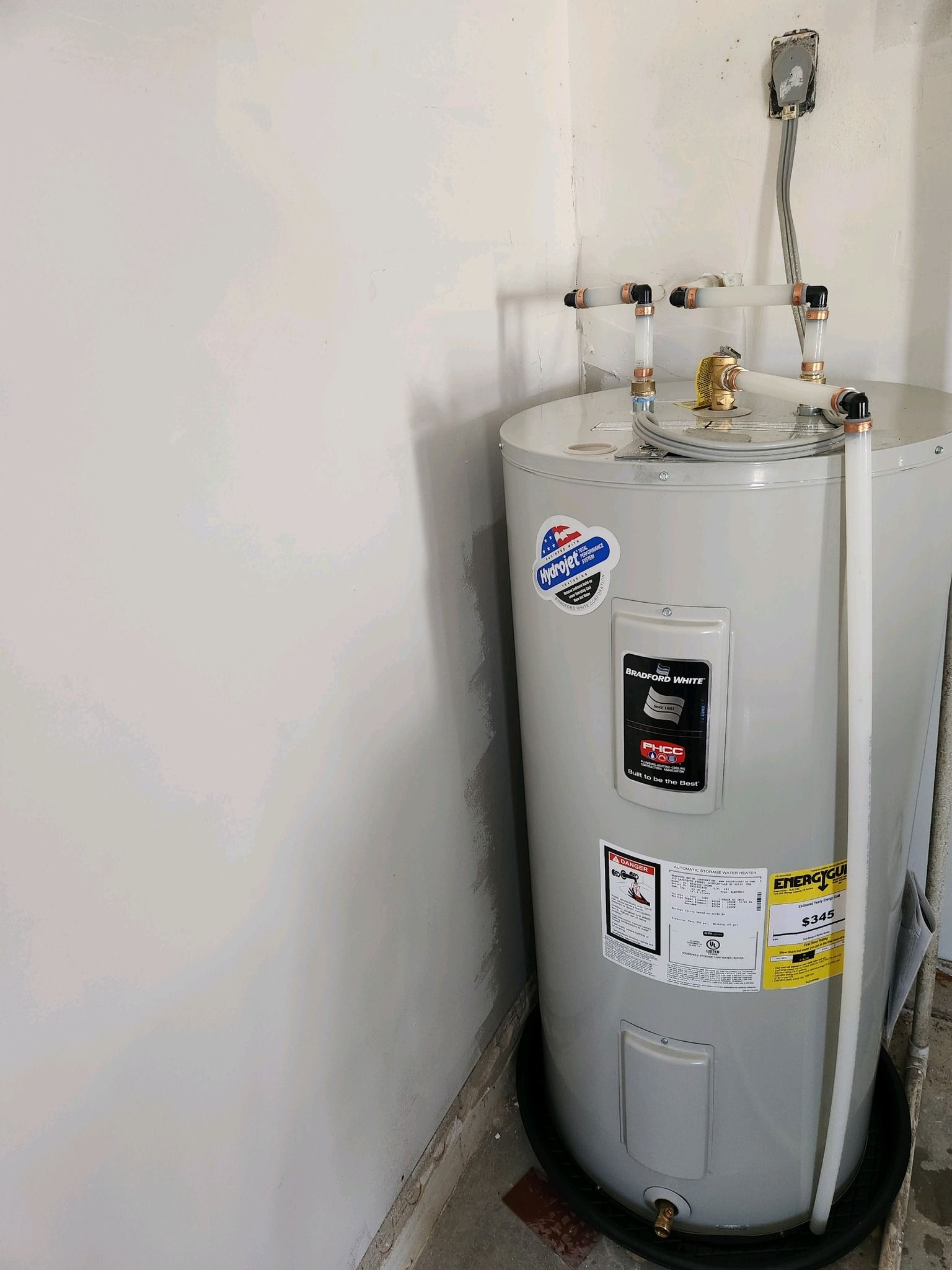 A gray residential water heater sits in a corner on a black drain pan, connected to white plumbing pipes overhead.