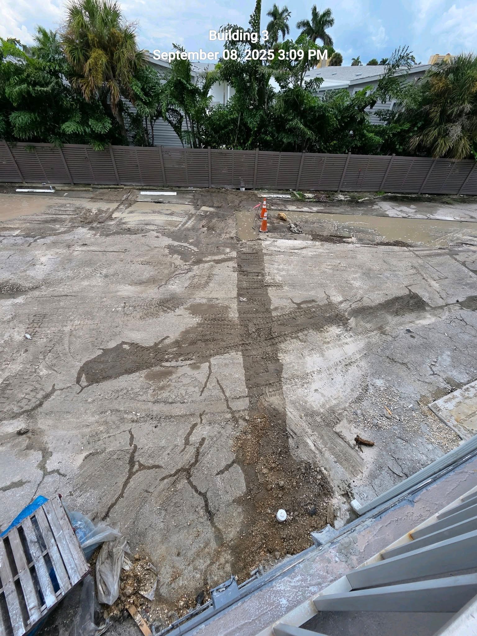 An outdoor concrete surface showing a cross-shaped trench or discoloration, with green trees and a fence in the background.