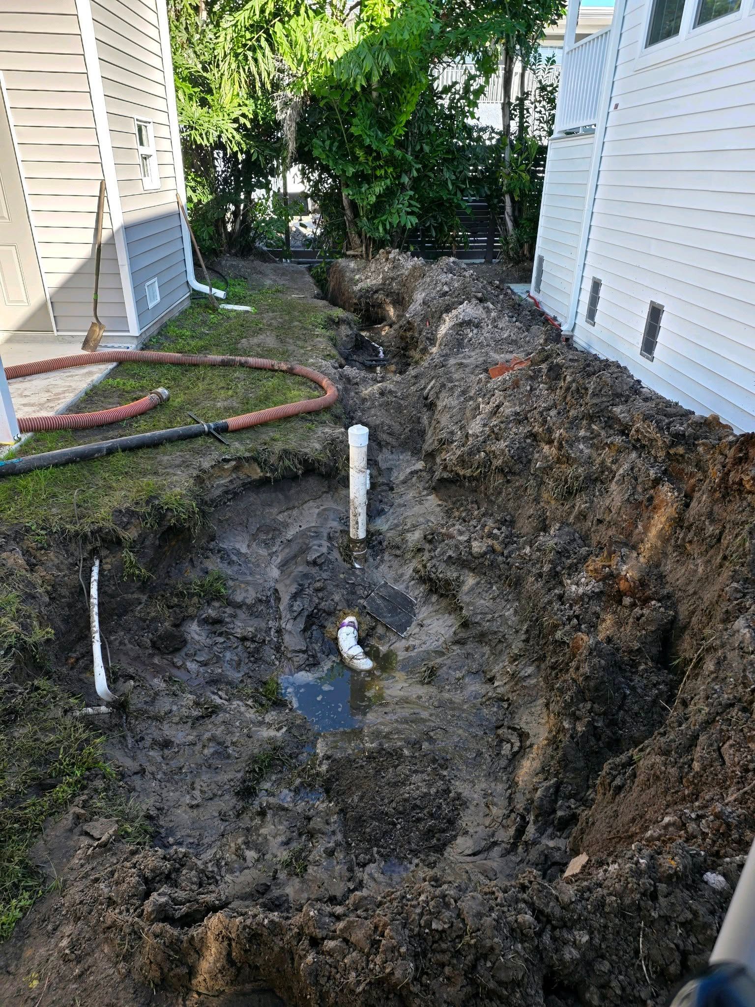 A muddy trench excavated between two houses with exposed plumbing pipes sticking out of the ground.