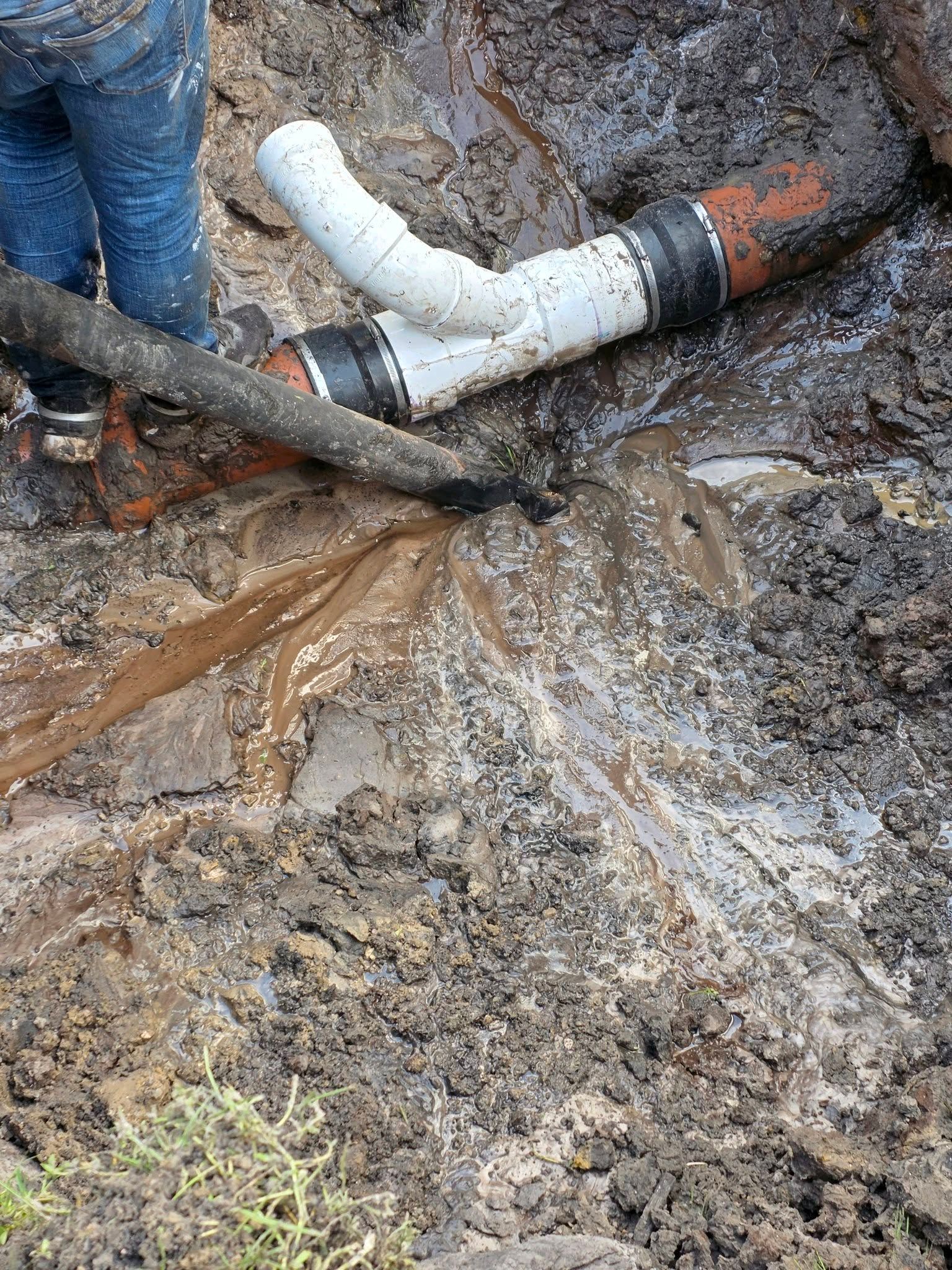 A worker repairs a residential sewer line in a muddy trench, installing a new PVC Y-fitting with rubber couplings.