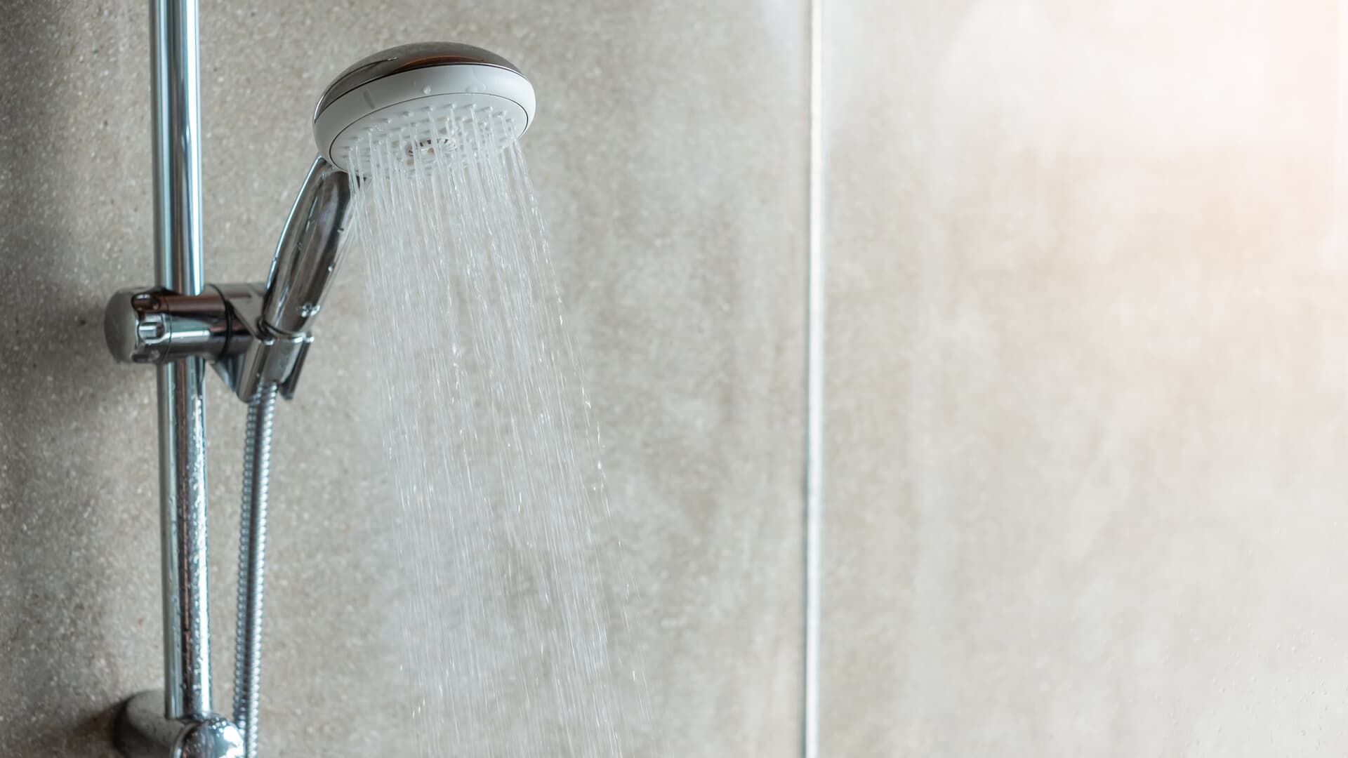 A chrome handheld shower head mounted on a wall bracket spraying water against a light grey tiled shower wall.