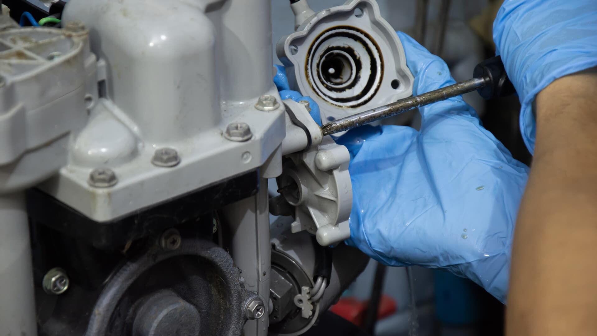A mechanic wearing blue gloves uses a screwdriver to remove a grey component from an engine assembly.