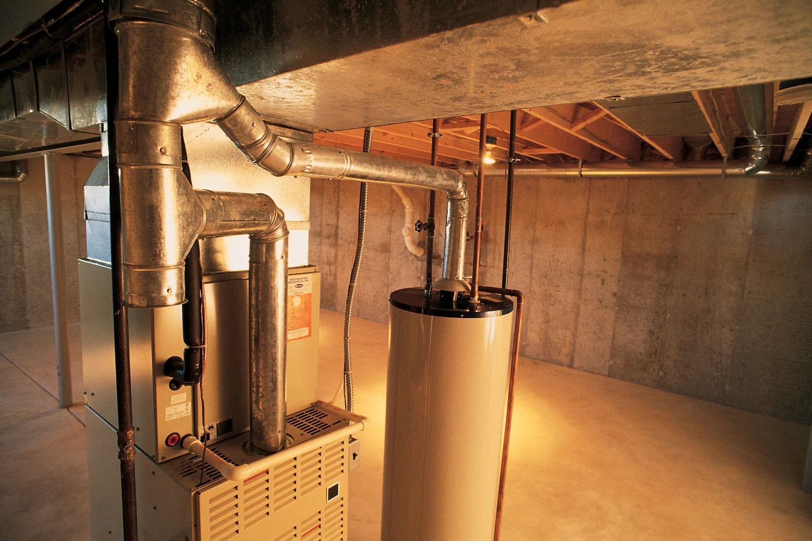 A water heater and furnace unit with metallic ventilation pipes in a basement with concrete walls and wooden ceiling joists.