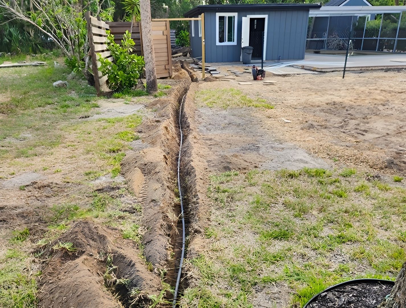 A narrow trench dug in a backyard lawn contains a black conduit pipe leading toward a small, gray shed.