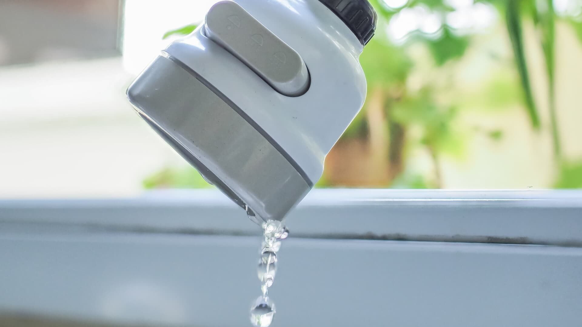 A gray, handheld faucet sprayer pouring a steady stream of water over a white surface with greenery in the background.
