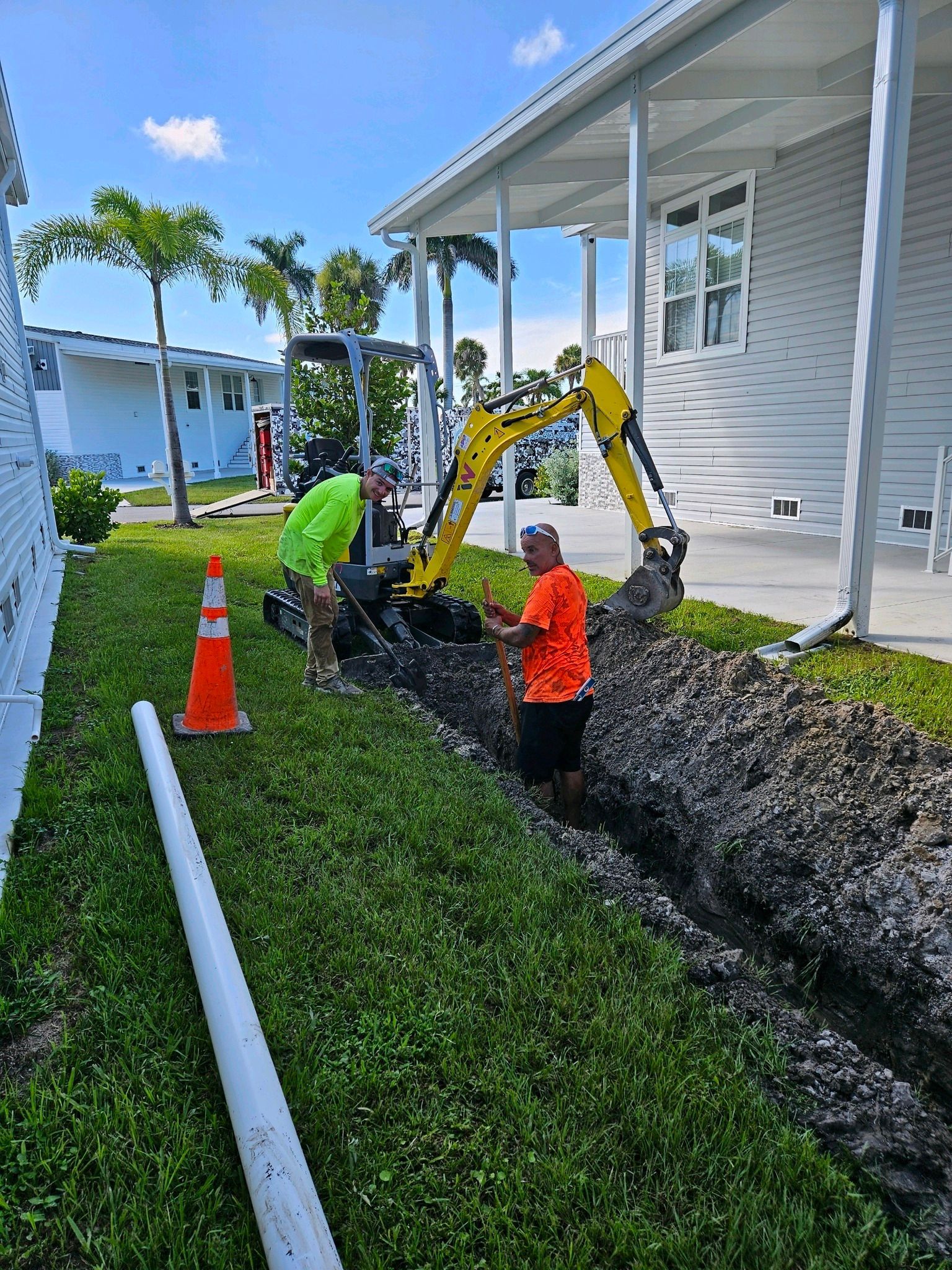 Two workers in high-visibility vests operate a small yellow excavator to dig a trench along a house.