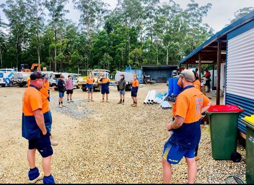 Construction Workers in Orange and Blue Uniforms Standing in a Circle Outdoors — Nambucca Plumbing in Kempsey, NSW