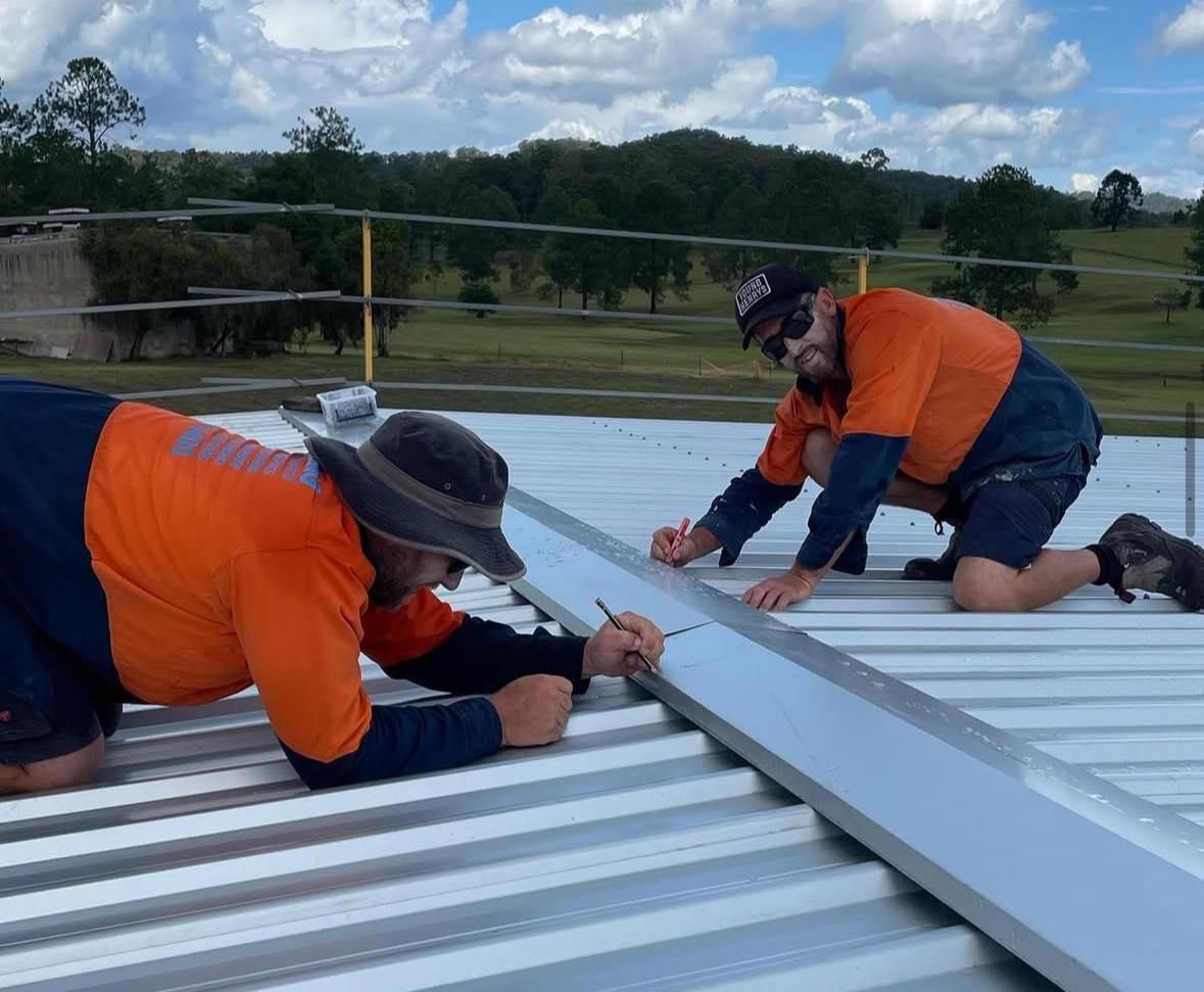 Two Workers in Orange Shirts Kneel on a Metal Roof — Nambucca Plumbing in South West Rocks, NSW