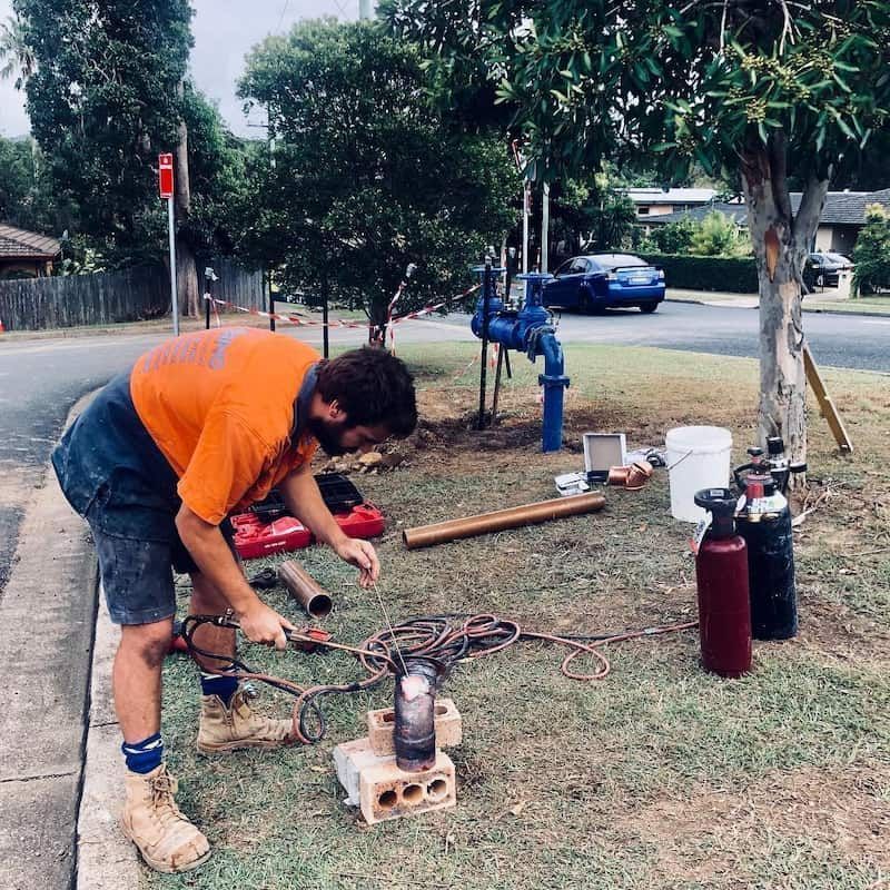 A Man in An Orange Shirt Is Working on A Pipe in The Grass — Nambucca Plumbing in Valla, NSW